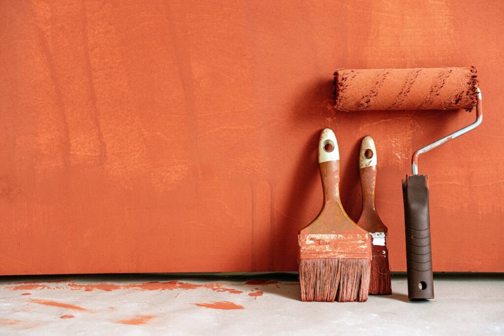 Close-up of paint rollers and brushes with orange paint against a painted wall.